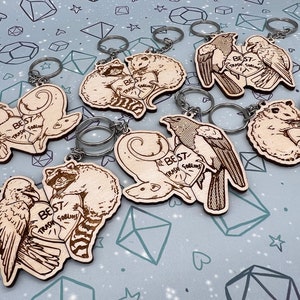 May include: Seven wooden keychains with different animal pairs. Each keychain features two animals holding a heart with the text 'Best Trash Goblins' on it. The animals include raccoons, birds, and possums. The keychains are on a light gray background with a pattern of white hearts and stars.
