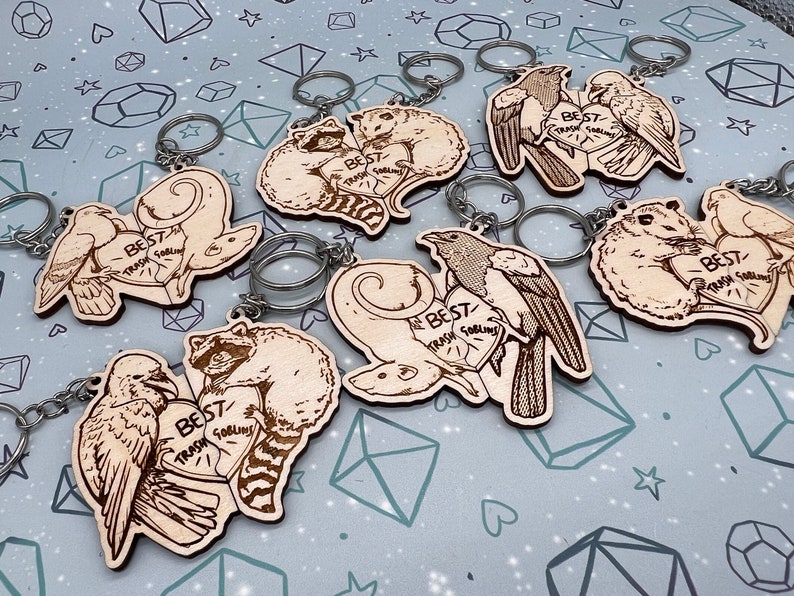 May include: Seven wooden keychains with different animal pairs. Each keychain features two animals holding a heart with the text 'Best Trash Goblins' on it. The animals include raccoons, birds, and possums. The keychains are on a light gray background with a pattern of white hearts and stars.