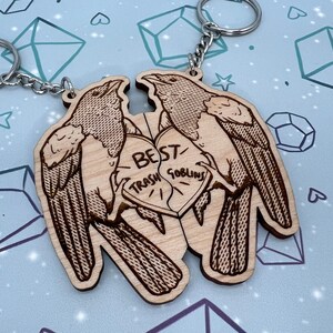 May include: Two wooden keychains shaped like birds with a heart in the middle. The heart is split in half and says 'Best Trash Goblins' on each half. The keychains are brown and have a detailed design.