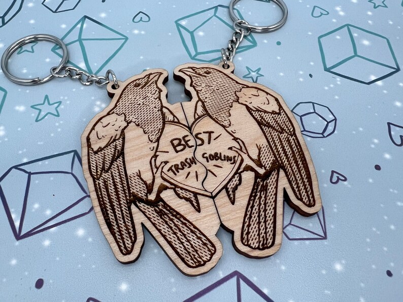 May include: Two wooden keychains shaped like birds with a heart in the middle. The heart is split in half and says 'Best Trash Goblins' on each half. The keychains are brown and have a detailed design.