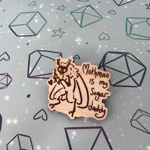 May include: A wooden pin with a cartoon mothman illustration and the text "Mothman is my Sugar Daddy".