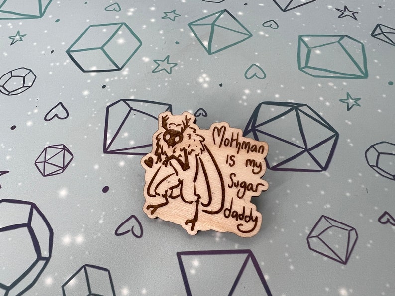 May include: A wooden pin with a cartoon mothman illustration and the text "Mothman is my Sugar Daddy".