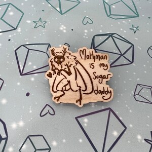 May include: A wooden pin with a cartoon mothman illustration and the text "Mothman is my Sugar Daddy".