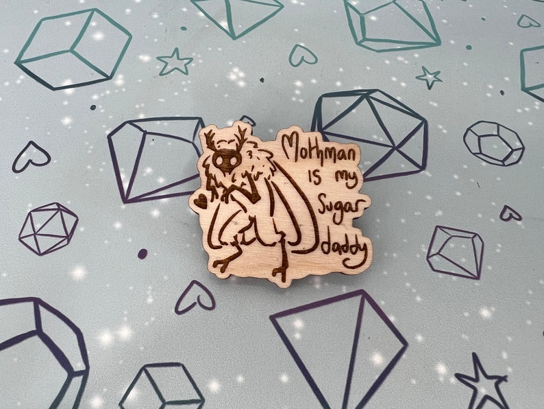May include: A wooden pin with a cartoon mothman illustration and the text "Mothman is my Sugar Daddy".
