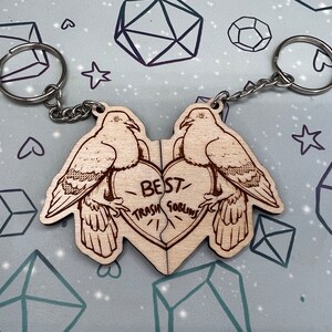 May include: Two wooden keychains shaped like doves with a heart in the middle. The heart is split in half and says 'Best Trash Goblins' on it. The keychains are connected by a silver chain.