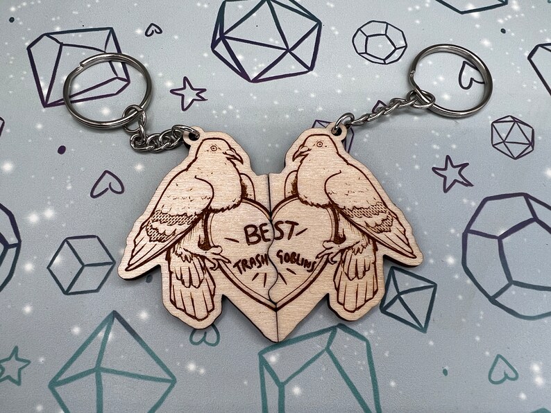 May include: Two wooden keychains shaped like doves with a heart in the middle. The heart is split in half and says 'Best Trash Goblins' on it. The keychains are connected by a silver chain.
