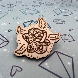 May include: A wooden pin with a detailed carving of two intertwined rings with eyes, surrounded by wings. The pin is on a light blue background with a pattern of hearts and diamonds.