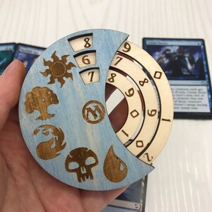 Könnte beinhalten: Ein drehbares Holzrad mit blauem Hintergrund und Symbolen für die fünf Farben von Mana in Magic: The Gathering. Das Rad hat die Zahlen 6, 7, 8 und 9.