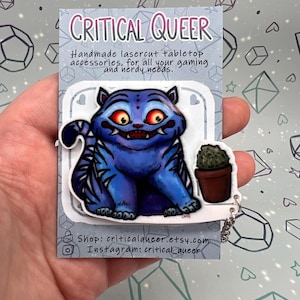 Peut inclure: Un personnage bleu et noir en forme de chat avec des yeux rouges et une petite plante en pot sur une carte blanche. La carte porte le texte "CRITICAL QUEER" et "Accessoires de table découpés au laser faits à la main, pour tous vos besoins de jeu et de geek."