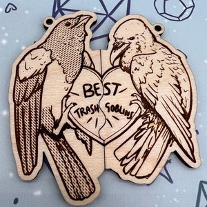 May include: Two wooden bird ornaments with a broken heart in the middle. The heart is inscribed with the words 'Best Trash Goblins'. The birds are facing each other and appear to be holding the heart together. The birds are detailed with black and brown lines and are on a light brown background.