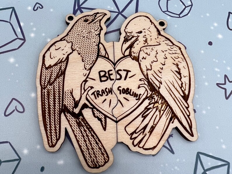 May include: Two wooden bird ornaments with a broken heart in the middle. The heart is inscribed with the words 'Best Trash Goblins'. The birds are facing each other and appear to be holding the heart together. The birds are detailed with black and brown lines and are on a light brown background.