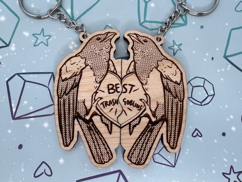 May include: Two wooden keychains shaped like birds holding a heart that says 'BEST TRASH GOBLINS' in a black font. The keychains are connected by a silver chain.