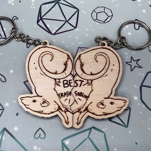 May include: A wooden keychain with two rats facing each other. The rats are carved in a heart shape and the text 'Best Trash Goblin' is written between them.