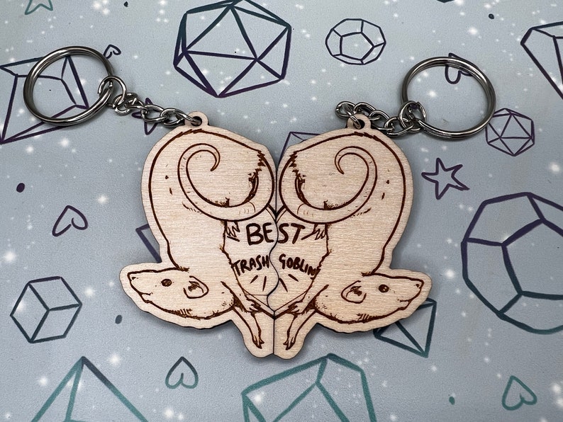 May include: A wooden keychain with two rats facing each other. The rats are carved in a heart shape and the text 'Best Trash Goblin' is written between them.