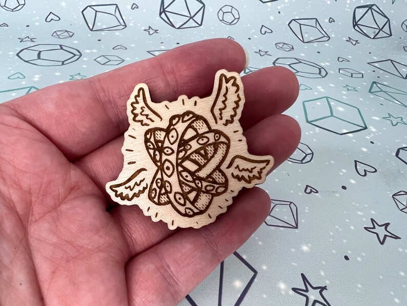 May include: A wooden cutout of a pair of eyes with wings, resembling a mythical creature. The eyes are detailed with intricate patterns and the wings are spread out.