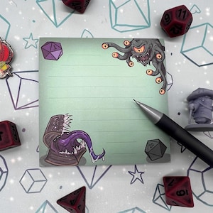 May include: A light green, lined sticky note with fantasy monster illustrations and a black pen. The note features a purple d20 die, a red and orange devil pin, and several red dice. A small gray figurine is also present.