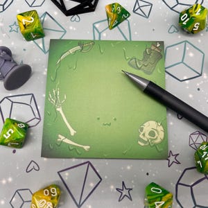 May include: A green square notepad with a skull, bones, and a sword illustration, next to a black pen, dice, and a figurine. The dice are green and yellow. The background is a light gray surface with geometric shapes.