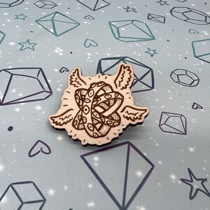 May include: A wooden pin with a detailed carving of a knot with eyes and wings. The knot is surrounded by a halo of light.