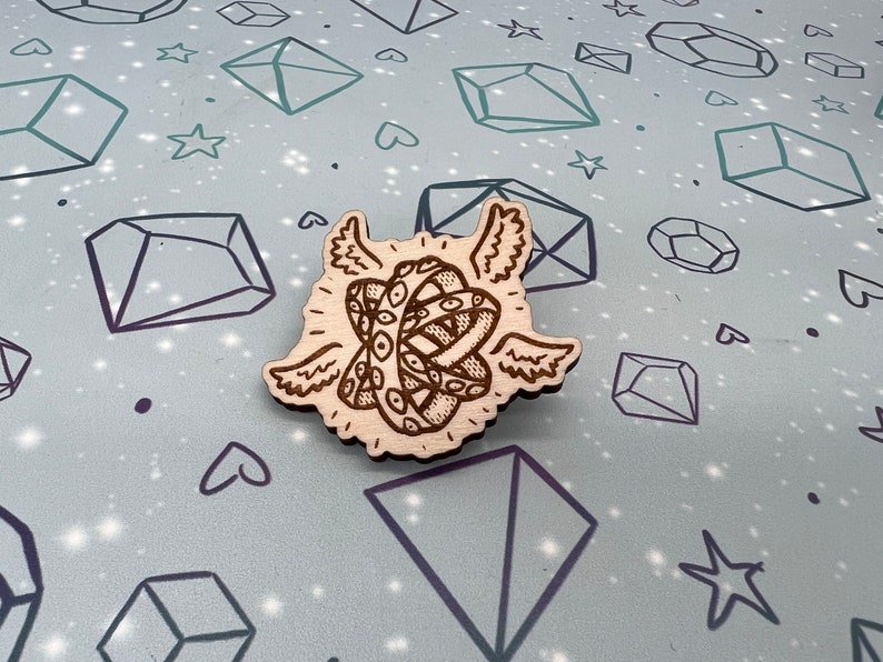 May include: A wooden pin with a detailed carving of a knot with eyes and wings. The knot is surrounded by a halo of light.