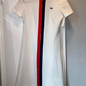 May include: A white short-sleeved dress featuring a central vertical zipper closure flanked by a red stripe on the left and a navy blue stripe on the right. The neckline is accented with matching red and navy blue trim, and a small blue crocodile logo is embroidered on the upper left chest.