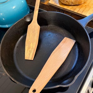 May include: A black cast iron skillet containing two wooden spatulas. One spatula is positioned vertically with a straight handle, while the other is placed diagonally and features a small circular hole at the end of its handle.