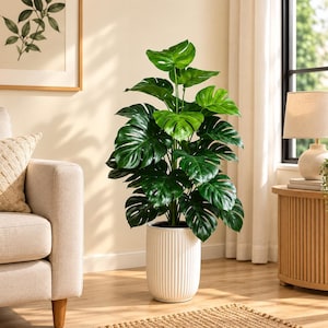 May include: A tall artificial monstera plant with large, glossy, dark green split leaves. The plant is housed in a cylindrical white planter featuring a vertical ribbed texture on the exterior, standing upright on a wooden floor surface.