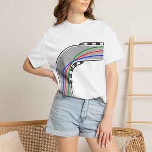 May include: A white short-sleeve t-shirt featuring a graphic of a gray curved racetrack with black and white checkered borders. The track design includes three distinct racing lines in red, green, and blue, creating a sharp cornering path.