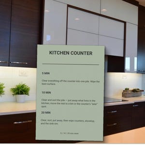 May include: A light green rectangular card with black text titled KITCHEN COUNTER. It lists cleaning tasks for 5, 10, and 20 minutes, including clearing, sorting, and wiping surfaces. The card is displayed against a modern kitchen background.