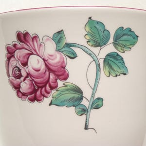 May include: A white ceramic cup with a floral design. The design features a large, detailed pink rose with green leaves and a teal stem. The rim of the cup is trimmed with a pink border.