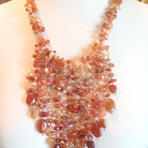 May include: A cascading necklace featuring a variety of orange, red, and cream-colored stones. The stones are strung together with gold-colored wire, creating a layered and textured effect. The necklace is displayed on a white mannequin.