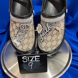 May include: A pair of black clogs featuring a tan and brown monogram pattern, embellished with silver rhinestones, a silver-tone double G logo, a white flower charm, a butterfly charm, and the word faith in script. A sign displays the text SIZE 9.