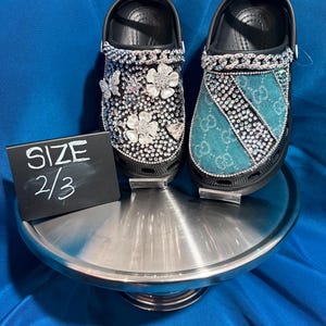 May include: A pair of black clogs decorated with rhinestones, silver chains, and flower charms. One clog features a teal fabric panel with a repeating logo pattern. A small black sign next to the shoes displays the white text SIZE 2/3.