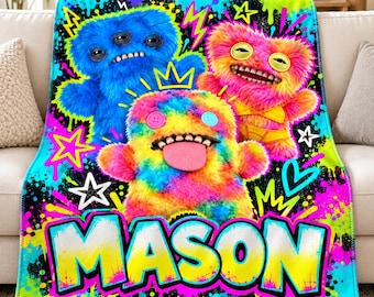 May include: A plush throw blanket featuring a vibrant, multi-colored graphic print of three fuzzy monsters. The design includes a blue, a pink, and a rainbow-colored monster with the name MASON printed in large, bold, yellow and white letters.