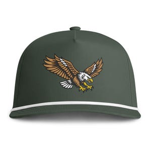 May include: A dark olive green baseball cap featuring a white cord trim across the base of the crown. The front panel displays a detailed graphic of a bald eagle in flight with brown wings, a white head, and yellow talons, captured in a diving pose.