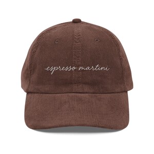 May include: A brown corduroy baseball cap featuring the text espresso martini embroidered in white cursive script across the front centre. The hat has a structured crown, a curved brim, and a button at the top, showcasing a textured fabric.
