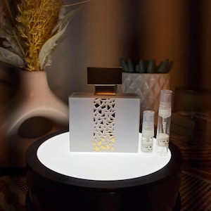May include: A rectangular white perfume bottle with a gold cap and a central vertical cutout featuring a geometric mosaic pattern. Two small clear sample spray vials are positioned to the right, each with a white label reading 2 ml M YLA.