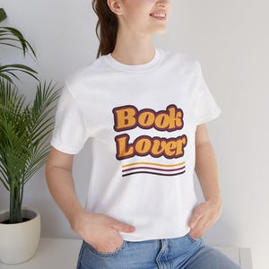 Puede incluir: Una camiseta blanca de manga corta con el texto "Book Lover" en una fuente de estilo retro. Las letras son amarillas con un borde granate oscuro. Debajo del texto hay tres líneas horizontales en colores amarillo, granate y marrón oscuro.