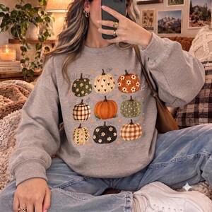 May include: A light grey crewneck sweatshirt featuring a graphic print of nine pumpkins in a grid layout. The pumpkins vary in colour and pattern, including plaid, floral, and heart designs, accented with small orange stars and hearts.