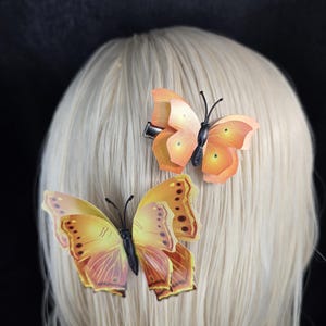 May include: Two hair clips featuring realistic butterfly designs. The top clip displays a bright orange butterfly with black antennae, while the bottom clip features a larger, yellow and orange butterfly with detailed wing patterns.
