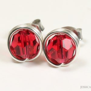 May include: A pair of stud earrings with bright red faceted crystal stones set in a silver-tone metal. The crystals are round and surrounded by a wire-wrapped design. The earrings are displayed on a white background.