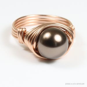 May include: A handcrafted ring featuring a large, dark gray pearl set in a copper-colored wire wrap. The wire is intricately woven around the pearl and forms the ring's band. The ring is displayed on a white background.