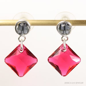 May include: A pair of dangle earrings featuring a square, faceted, hot pink crystal pendant. The earrings have a small, gray bead at the top, connected by silver-colored wire. The earrings are displayed on a gold-colored bar.