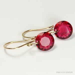 May include: A pair of gold-colored drop earrings featuring large, faceted, round, ruby-red gemstones. The earrings have a simple, elegant design with a hook closure. The gemstones are the focal point, catching the light and adding a pop of color.