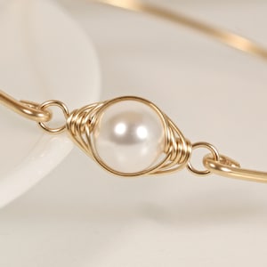 Puede incluir: Un brazalete de oro con una sola perla blanca envuelta en alambre dorado.