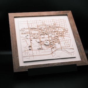 May include: A square, framed 3D map featuring a white, raised relief city layout with brown engraved street lines. The map is encased in a dark, textured wooden frame with a white mat border, presenting a detailed architectural model of a city grid.