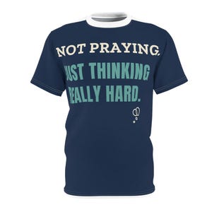 May include: A navy blue short-sleeve t-shirt with a white crew neckline. The front features the text "NOT PRAYING, JUST THINKING REALLY HARD." in white and light blue lettering, accompanied by a small white graphic of thought bubbles.