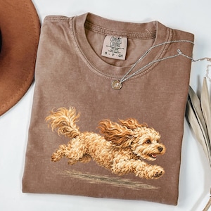 May include: A brown short-sleeved t-shirt featuring a graphic of a running golden-colored curly-haired dog. The shirt has a Comfort Colors brand label at the inner neckline and a silver-toned chain necklace with a small circular pendant.