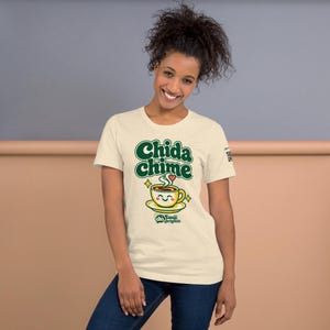 Smiling woman with curly hair wearing a Heather Natural t-shirt printed with green retro letters reading Chida chime above a happy yellow coffee cup, against a peach and gray wall.