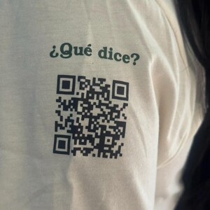 Close-up of the t-shirt sleeve printed with "¿Qué dice?" in green and a black QR code that opens emojijerigonza.com.