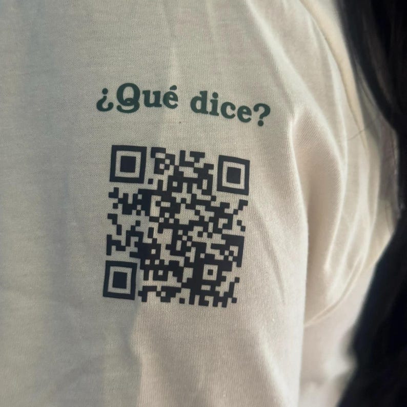 Close-up of the t-shirt sleeve printed with "¿Qué dice?" in green and a black QR code that opens emojijerigonza.com.